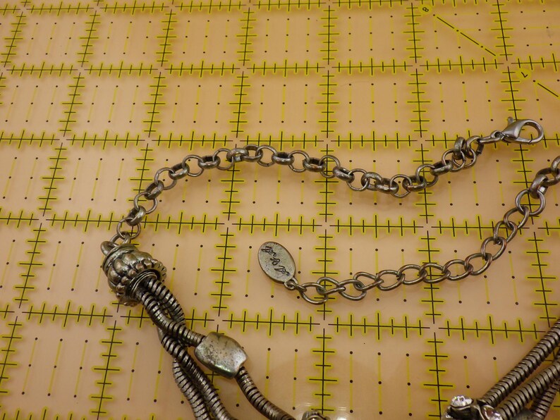 May include: A silver-toned multi-strand necklace featuring flexible snake chains, a decorative ribbed bead, and a small oval metal tag with engraved text. The necklace includes a lobster claw clasp and an adjustable chain link extender.
