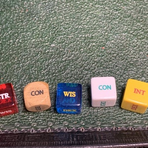 May include: Five dice with different colors and labels. The dice are red, brown, blue, white, and yellow. The labels on the dice are STR DEX, CON DEX, WIS DEX, CON DEX, and INT DEX.