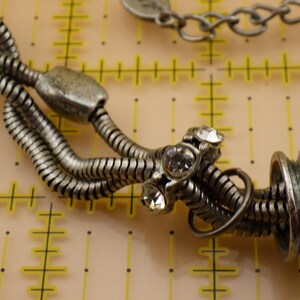 May include: A silver-toned multi-strand snake chain bracelet featuring a central metal bead set with three clear round rhinestones and a larger cylindrical textured bead. The bracelet is positioned over a yellow grid mat with inch markings.