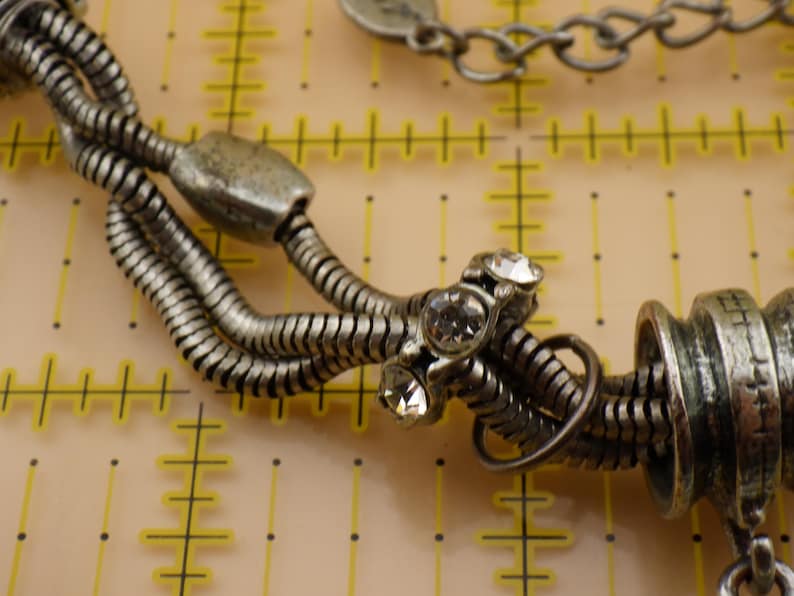 May include: A silver-toned multi-strand snake chain bracelet featuring a central metal bead set with three clear round rhinestones and a larger cylindrical textured bead. The bracelet is positioned over a yellow grid mat with inch markings.