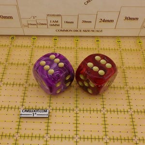 May include: Two six-sided dice, one purple and one red, both featuring a marbled finish and cream-colored pips. The dice are positioned on a yellow grid mat with a scale reference showing a 1-inch marker labeled CAVELOOT.COM and common dice sizes.