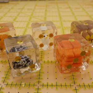May include: Five clear dice with different colorful objects inside. The dice are made of clear plastic and have a gold-colored border. The objects inside the dice are a white bird, a brown and black rock, a small orange and gold object, and two brown and gold objects.