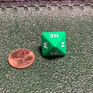May include: A green six-sided die with white lettering. The sides of the die are labeled with the directions: N, NE, E, SE, S, and W.