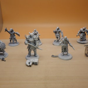May include: A collection of unpainted miniature fantasy figures, including warriors and creatures, with various weapons and armor. A six-sided die with a logo is also present. The figures are on a light brown surface.