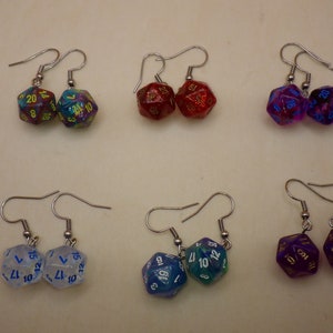 May include: A collection of six pairs of D20 dice earrings. Each earring features a dice with numbers visible, attached to a silver-colored fish hook. The dice are in various colors, including red, blue, purple, and clear.