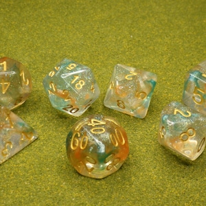 May include: Set of seven polyhedral dice with clear resin bodies, infused with gold glitter and swirls of orange and teal. The numbers are painted in gold. The dice are arranged on a green surface.