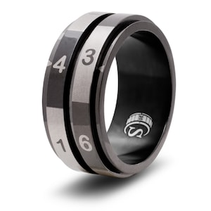 May include: A black and silver spinning ring with the numbers 1, 3, 4, and 6 engraved on the sides. The ring has a textured surface and a logo on the inside.