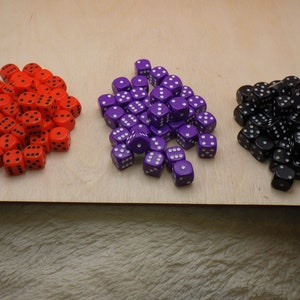 May include: Three piles of dice in red, purple, and black. Each die has white dots. The dice are on a light wood surface. The dice are for board games and tabletop games.