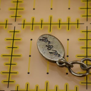 May include: An oval-shaped metallic jewelry tag with a dark, textured finish and an engraved signature, attached to a metal chain link. The tag is positioned over a yellow grid background with markings that indicate a measurement of 0.5 inches.