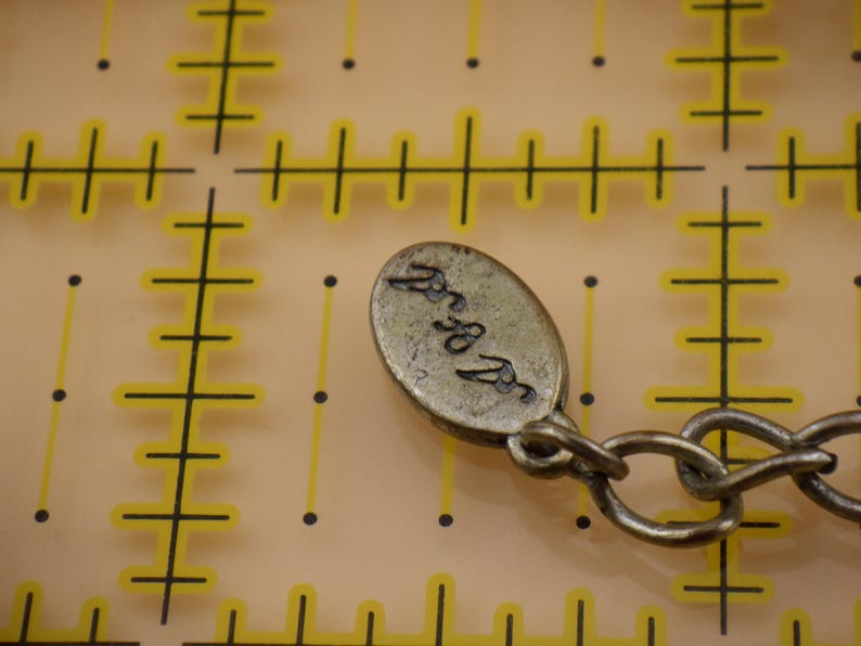 May include: An oval-shaped metallic jewelry tag with a dark, textured finish and an engraved signature, attached to a metal chain link. The tag is positioned over a yellow grid background with markings that indicate a measurement of 0.5 inches.