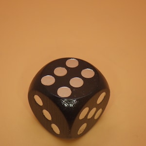 May include: A black wooden dice with white dots. The dice is showing the number five.
