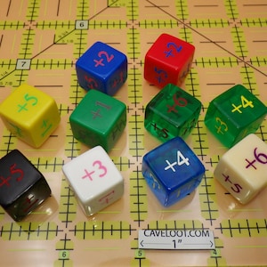 May include: A collection of ten multi-colored dice with plus signs and numbers, including blue, red, green, yellow, black, and white. The dice are arranged on a grid with the text "CAVELOOT.COM" and a 1-inch ruler.