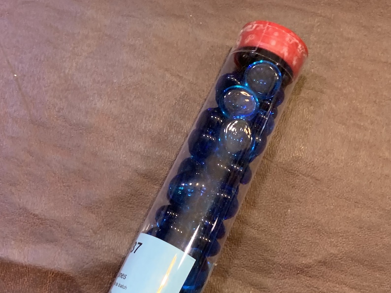 May include: A clear glass tube filled with small, round, blue glass marbles. The tube has a red lid and a white label with the number "37" on it.