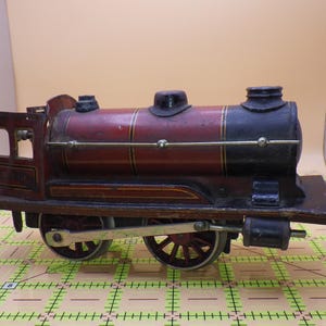 May include: Vintage toy train with a maroon and black color scheme. The train features a cylindrical boiler, a detailed cab, and spoked wheels. The model has gold accents and appears to be made of metal.