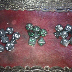 May include: A collection of black dice with white numbers and green accents. The dice are scattered on a red leather surface.