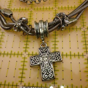 May include: A silver-toned necklace featuring a multi-strand snake chain with decorative metal beads and clear rhinestones. A central cross pendant with intricate scrollwork and embedded clear rhinestones hangs from the chain, positioned over a grid.