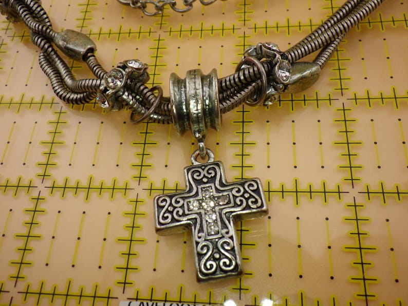 May include: A silver-toned necklace featuring a multi-strand snake chain with decorative metal beads and clear rhinestones. A central cross pendant with intricate scrollwork and embedded clear rhinestones hangs from the chain, positioned over a grid.