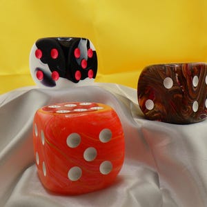 May include: Three large dice with different patterns. The first die is black and white with red dots, the second is orange with white dots, and the third is brown with white dots.