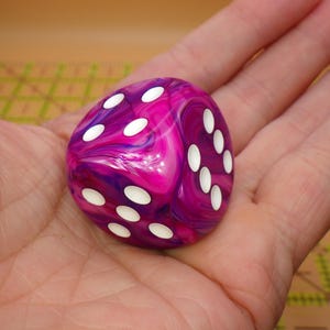 May include: A six-sided die featuring a marbled pattern of vibrant magenta, purple, and white swirls. The die has rounded corners and smooth surfaces, with recessed circular pips colored in white, clearly displaying the numbers four and six.
