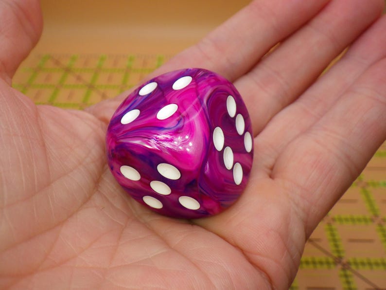 May include: A six-sided die featuring a marbled pattern of vibrant magenta, purple, and white swirls. The die has rounded corners and smooth surfaces, with recessed circular pips colored in white, clearly displaying the numbers four and six.