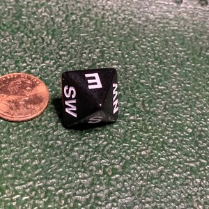 May include: A black, six-sided dice with white letters on each side. The letters are E, S, W, N, NW, and SW.