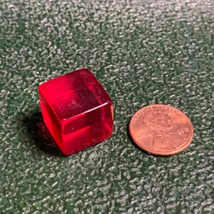May include: A small, red, transparent cube next to a US penny.
