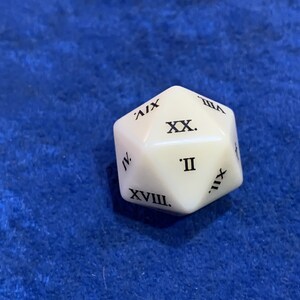 May include: A white twenty-sided die with black Roman numerals. The die is resting on a blue surface. The numerals are clearly visible, indicating the numbers for a tabletop game.