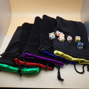 May include: Six colorful dice with iridescent finishes are displayed on a table, resting inside five black velvet drawstring bags with satin trim in various colors. The bags are arranged in a stack, with the dice visible in the top bag.
