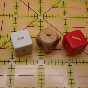 May include: Three dice with different colors and text. A white die reads "STAFF", a wooden die reads "ROD", and a red die reads "SCEPTRE". The dice are on a yellow grid with measurements in inches.