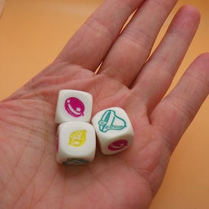 May include: Three white dice with different colored symbols. One die has a pink and green planet, one has a yellow lemon, and one has a green bell.