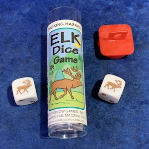May include: A clear plastic tube with a red lid contains two white dice with brown elk images. The tube has a label that reads "CHOKING HAZARD ELK Dice Game" and includes the company name and address.