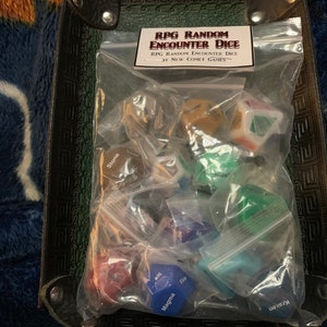 May include: A clear plastic bag filled with various colored dice. The dice are labeled with words like "Stone", "Ice", "Magma", "Kraken", and "Fire". The bag is labeled "RPG Random Encounter Dice" and "By New Comet Games".