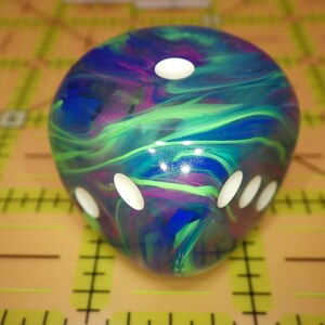 May include: A six-sided die featuring a swirling marbled pattern of deep blue, vibrant purple, and bright green colors. The die has rounded edges and smooth surfaces with recessed white circular pips indicating the numerical values on each face.
