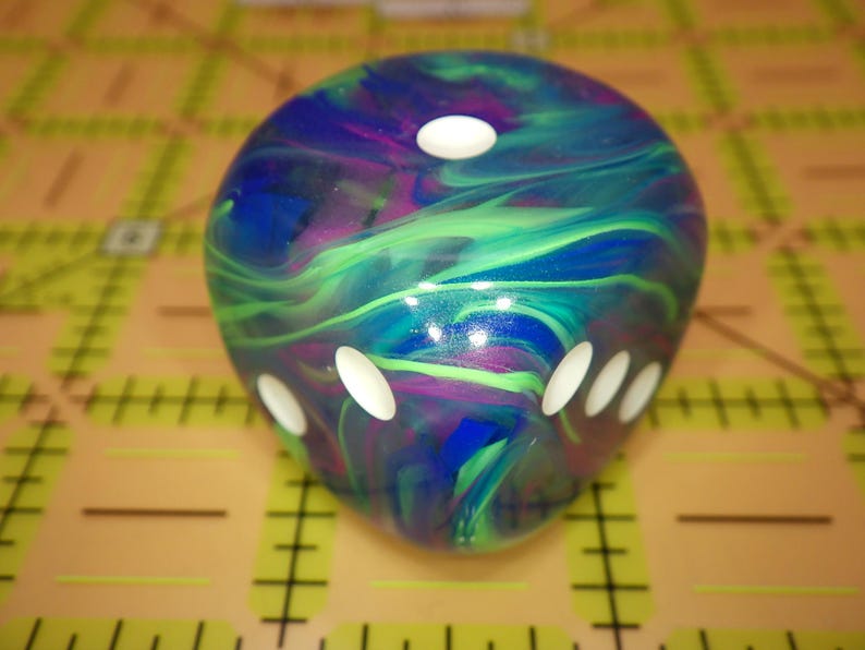 May include: A six-sided die featuring a swirling marbled pattern of deep blue, vibrant purple, and bright green colors. The die has rounded edges and smooth surfaces with recessed white circular pips indicating the numerical values on each face.