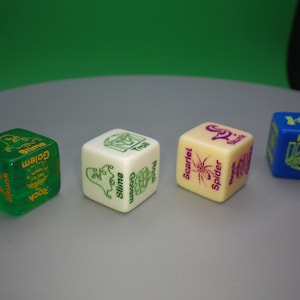 May include: Four dice with different colors and designs. The dice are green, white, cream, and blue. The green die has the words "Gollum", "Slime", "Rock", and "Shun" on it. The white die has the words "Gollum", "Slime", "Troll", and "Rock" on it. The cream die has the words "Scarlet Spider", "Rock", "Troll", and "Slime" on it. The blue die has the words "Troll", "Slime", "Rock", and "Gollum" on it.