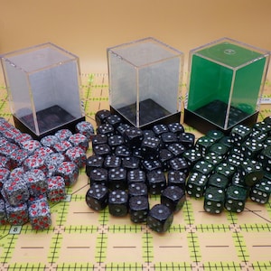May include: Three sets of dice with clear acrylic display boxes. One set is red and gray with red pips, another is black with white pips, and the third is green with white pips. The dice are arranged on a yellow grid.