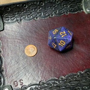 May include: A purple and black speckled dice with gold numbers 2, 11, 10, 12, 15, and 8 on a black leather dice tray with a brown felt interior. The tray has a decorative border and the letters "GS" are visible in the bottom left corner.
