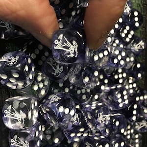 May include: A collection of translucent purple dice with white pips and a white graphic design. The dice are being held by a hand. The dice are for tabletop games.