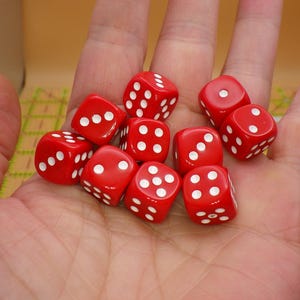 May include: A handful of red dice with white dots. The dice are a vibrant red color and have white dots indicating the numbers. The dice are held in an open hand, suggesting they are ready to be rolled for a game.