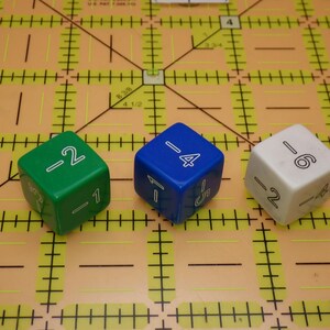 May include: Three dice with negative numbers and other markings are displayed on a yellow cutting mat. The dice are green, blue, and white. The green die shows -2 and -1. The blue die shows -4, 1, and 5. The white die shows -6 and -2.