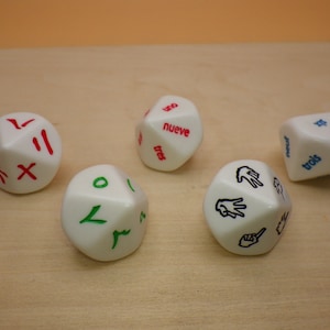 May include: Five white dice with different colored and black markings. The dice have the numbers one, two, three, four, five, six, seven, eight, nine, ten, eleven, and twelve written on them. The dice are on a light brown wooden surface.