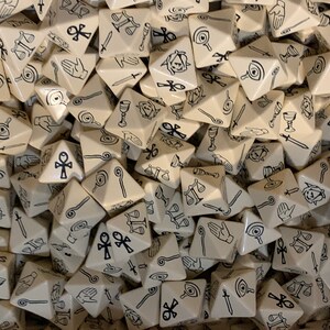 May include: A pile of beige triangular dice with black symbols. The symbols include an eye, a hand, a staff, a chalice, a sword, a scale, and an ankh.