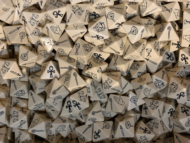 May include: A pile of beige triangular dice with black symbols. The symbols include an eye, a hand, a staff, a chalice, a sword, a scale, and an ankh.