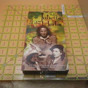 May include: A VHS tape titled "Juliette of the Herbs" featuring a vintage book cover design. The cover displays a portrait of a woman and the text "The most important thing we can teach children is love for animals." The tape is on a clear ruler.