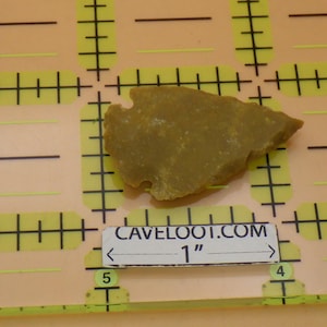 May include: A tan-colored, irregularly shaped stone artifact, possibly an arrowhead, is displayed on a yellow grid. The artifact is approximately 1 inch in length. The text "CAVELOOT.COM" and "1" are visible.
