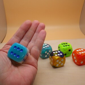 May include: Five dice with different colors and patterns. The dice are blue, green, yellow, and orange. The dice are on a light brown surface.