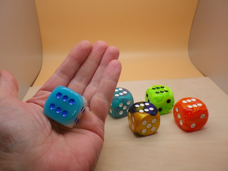 May include: Five dice with different colors and patterns. The dice are blue, green, yellow, and orange. The dice are on a light brown surface.