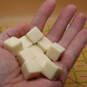May include: A hand holds a collection of small, square, ivory-colored game tiles. The tiles are smooth and uniform in shape, with a slightly rounded edge. The background is a warm, neutral tone, suggesting a tabletop setting.