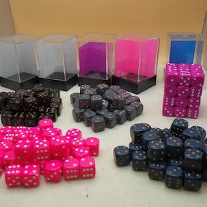 May include: A collection of dice in various colors, including black, pink, and blue, with gold dots. The dice are displayed in clear plastic containers and on a white surface.