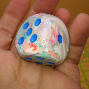 May include: A six-sided die with a marbled, multi-colored surface featuring swirls of white, pink, and teal. The die has rounded edges and is marked with bright blue circular pips on its faces, showing the numbers three and one clearly.